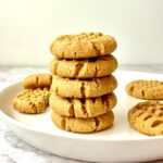 A stack of peanut butter cookies on a plate, surrounded by more peanut butter cookies.