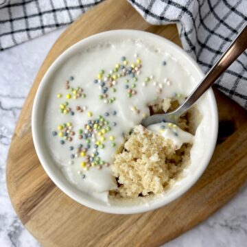 A spoon in a vegan vanilla mug cake topped with frosting and sprinkles.