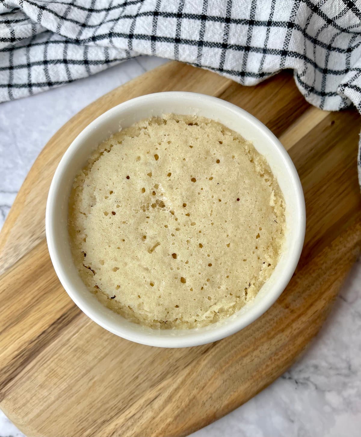 A vanilla mug cake in a ramekin sitting on a wooden cutting board.