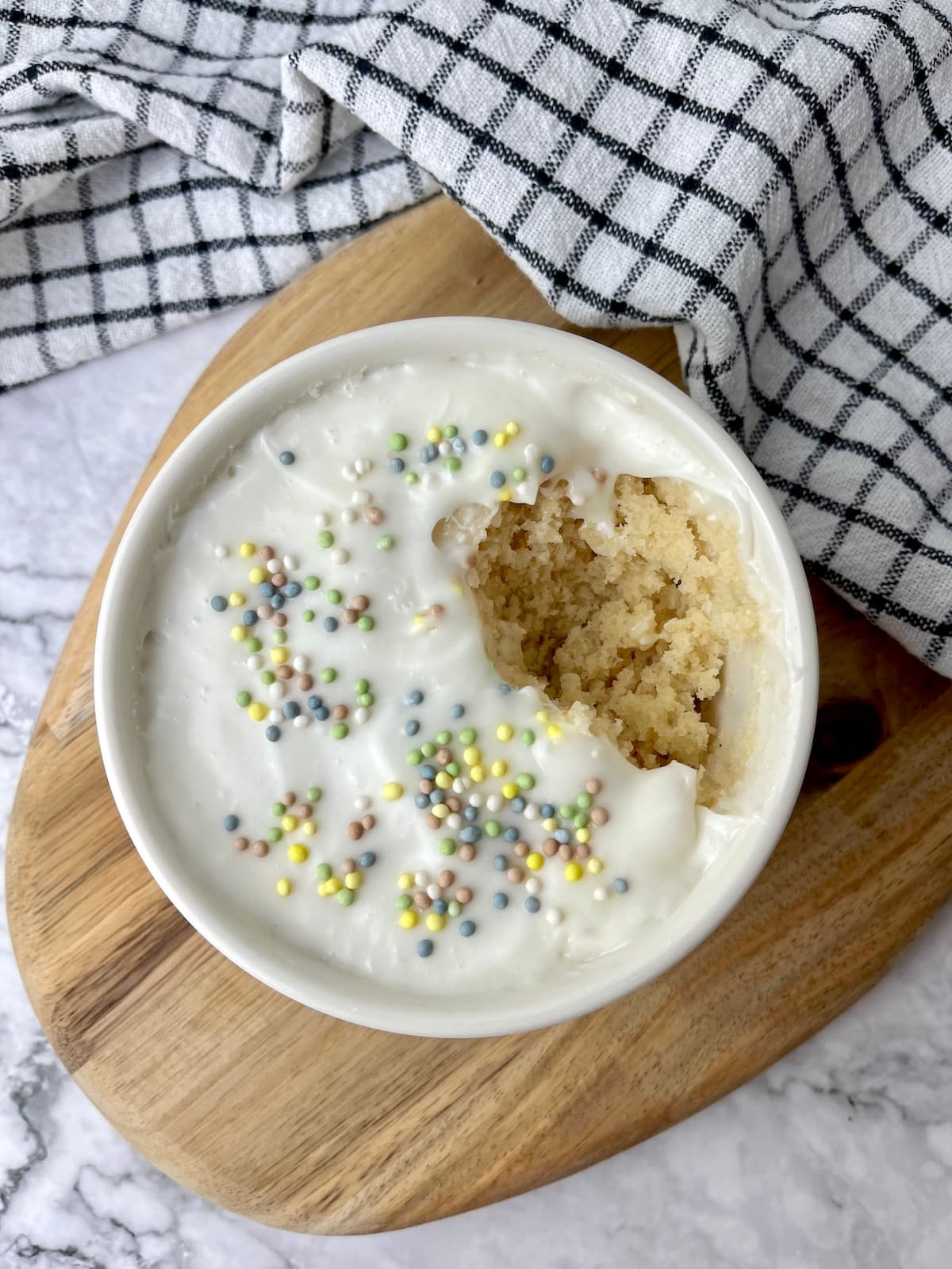 A vegan vanilla mug cake topped with frosting and sprinkles, with a bite taken out of it.
