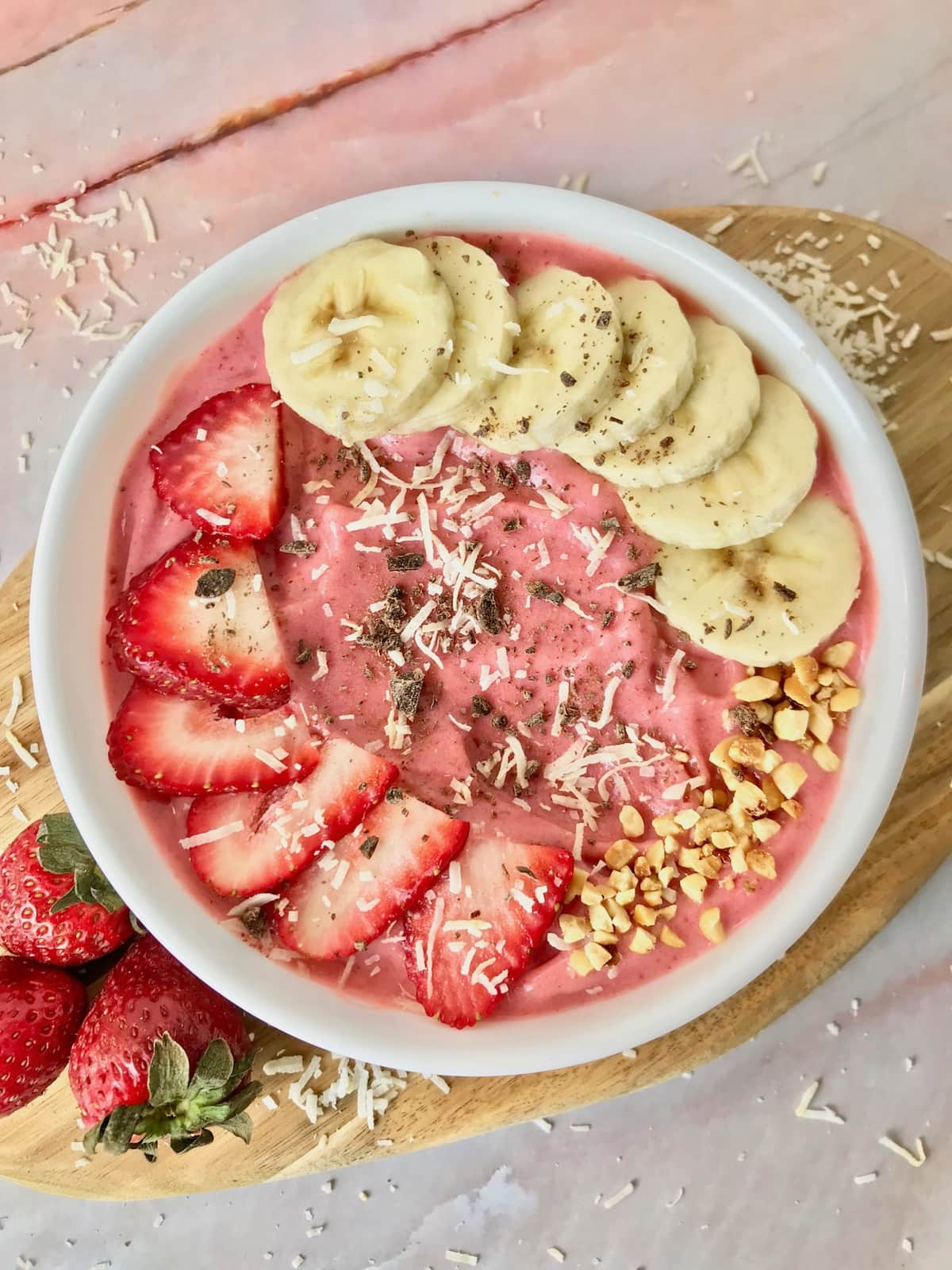 A strawberry banana smoothie bowl topped with banana slices, strawberry slices, coconut, peanuts, and chocolate.